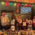 Group of people seated and standing around a restaurant-style table holding illustrated portraits, with string lights and framed artwork in the background during a fellowship social event.