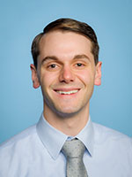 Professional headshot of an individual wearing a light blue dress shirt and a light gray tie against a solid blue background.