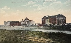 Colorized photograph. Foreground, there are railroad tracks running across grassy land, behind which is a canal. Far side stands a group of large brick decorative Center-left is a lighter-colored building with a dome-shaped roof.