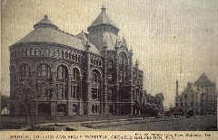 Black and white photograph of a large, ornate brick building with arched windows and decorative stonework (right) on the left is a building with a domed roof.