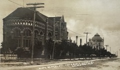 On the left is a large, ornate brick building with arched windows and decorative stonework, Utility poles with multiple crossbars and overhead wires run along the street. Background (right) there is another building with a large dome-like roof.