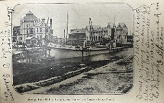 A black‑and‑white photograph shows a series of ornate buildings and a workboat dredging on a canal.