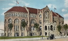 Colorized photograph of a large, ornate brick Romanesque Revival style building with arched windows and decorative stonework, with a central staircase. A car is parked on the roadside. Flags are flying either side of the roof.
