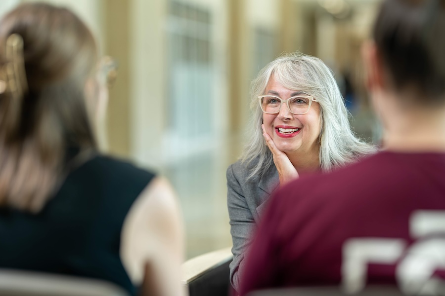 A person with gray hair and glasses sits facing two other people during an indoor conversation, with chairs and a softly lit hallway visible in the background