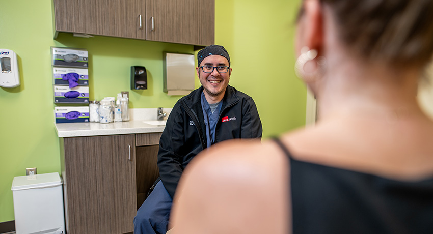 Clinician seated in an exam room faces a patient in the foreground, with cabinets, a sink, and medical supplies visible behind the clinician.