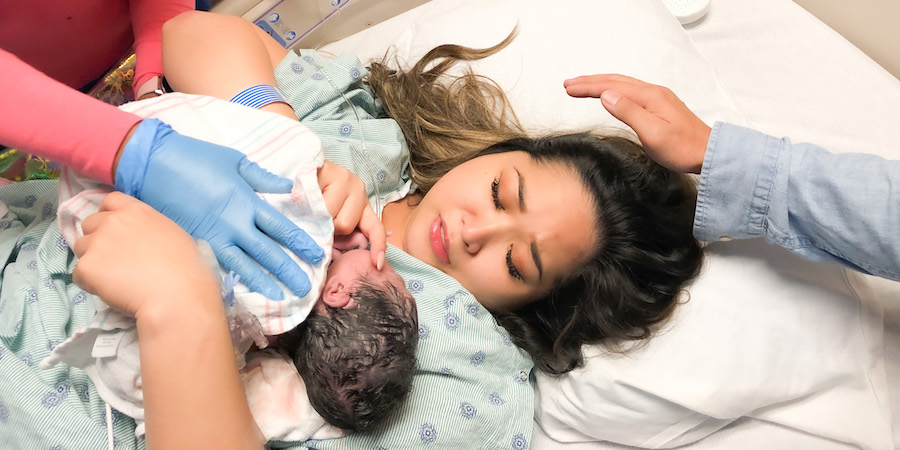brunette female patient in hospital gown laying in bed with baby on her chest with a blanket around baby and a nurses gloved hand on baby's back. another hand is near mom's head for support