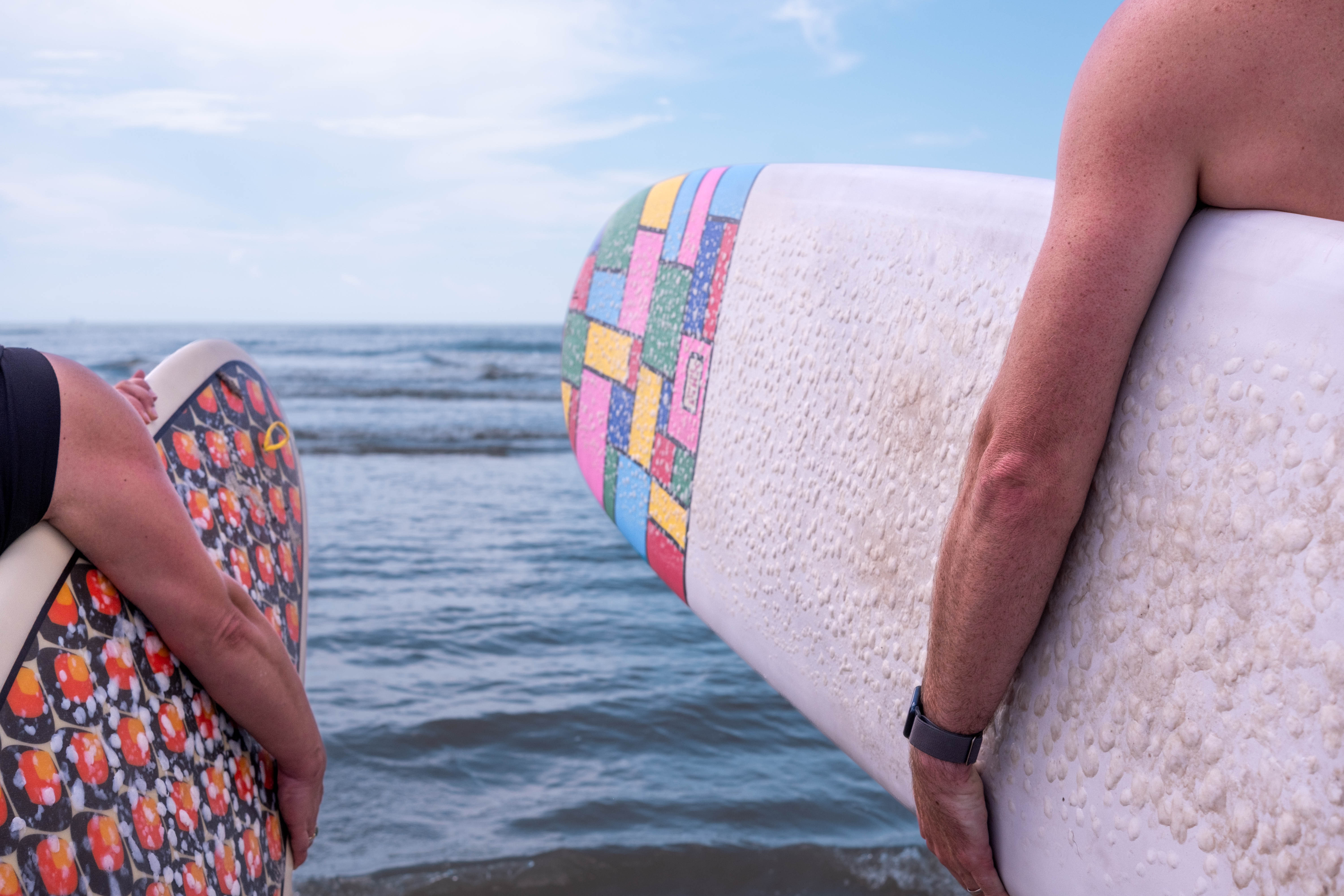 As an island, Galveston offers its residents many opportunities for water-based extracurriculars that encourage physical activity, like surfing. Two people stand at the shoreline holding surfboards, facing the water. One surfboard has a colorful patchwork design, and the other has a patterned graphic.
