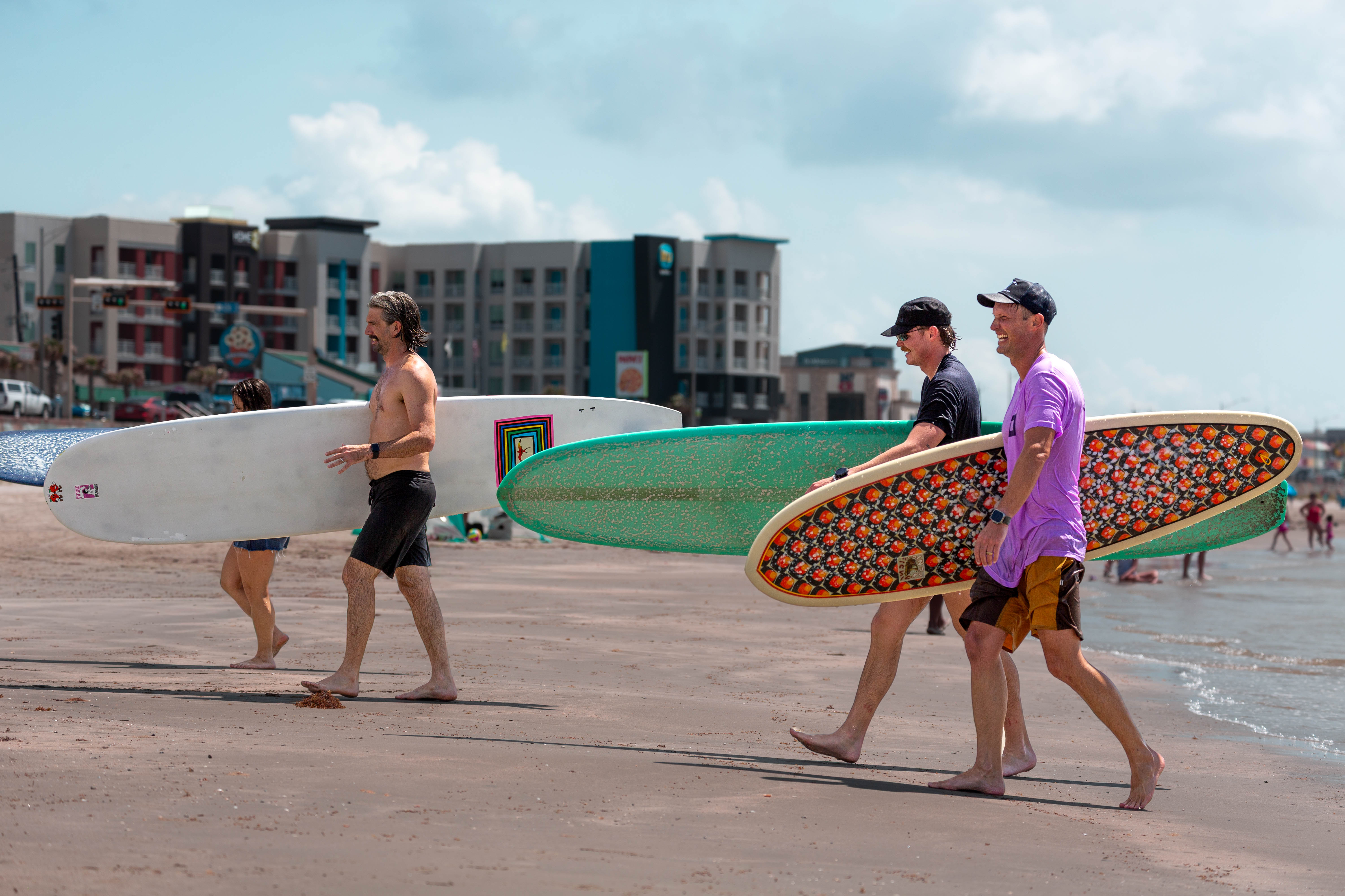 Activities like surfing also foster social connections within the community, an important part of the lifestyle promoted by the Blue Zones Project. Four people carry surfboards while walking along the shoreline. Buildings and beachgoers are visible in the background near the water.