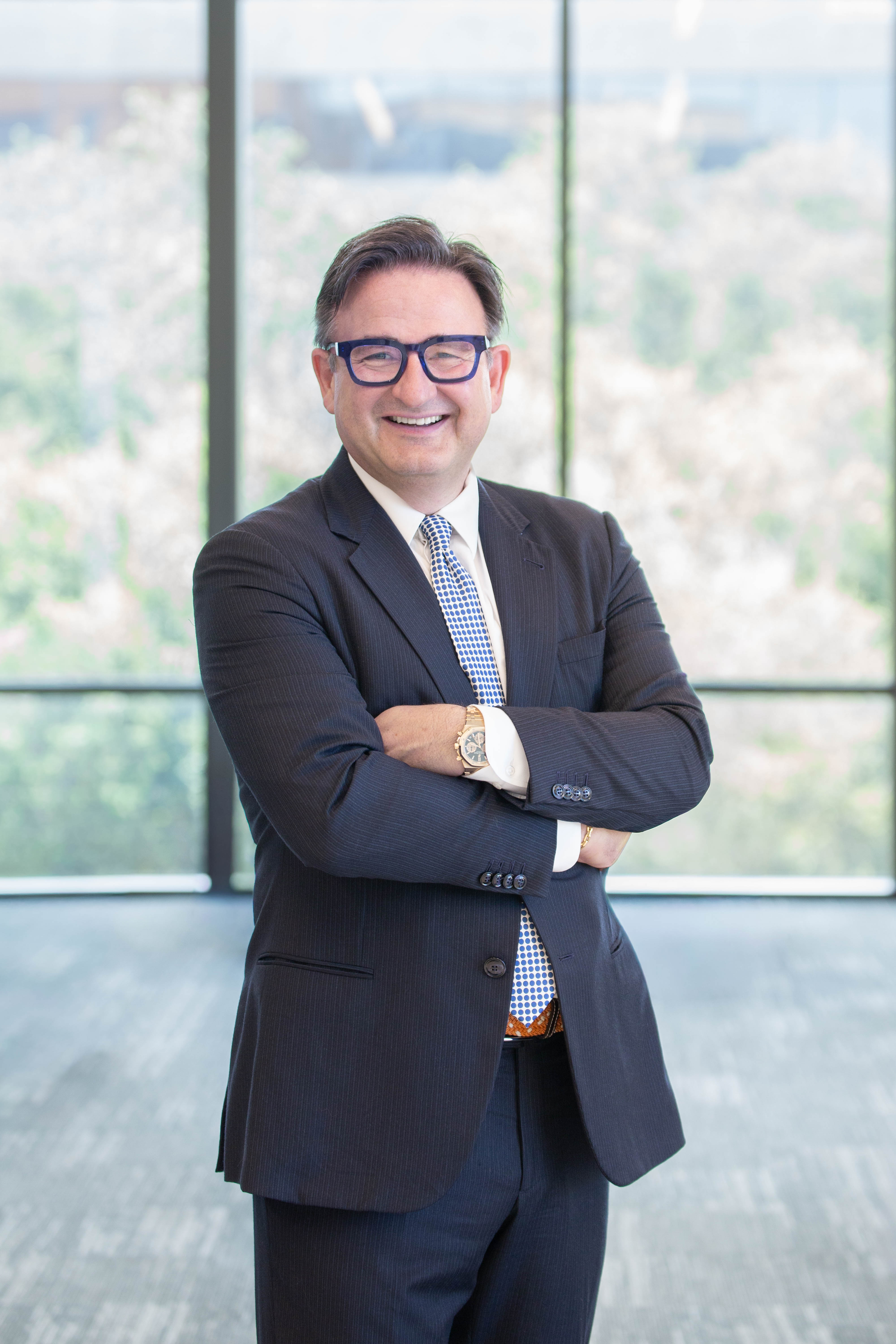Dr. Jochen Reiser, President of UTMB and CEO of the UTMB Health System. Person wearing a dark suit, patterned tie, and glasses stands with arms crossed in front of large windows with a blurred outdoor view.