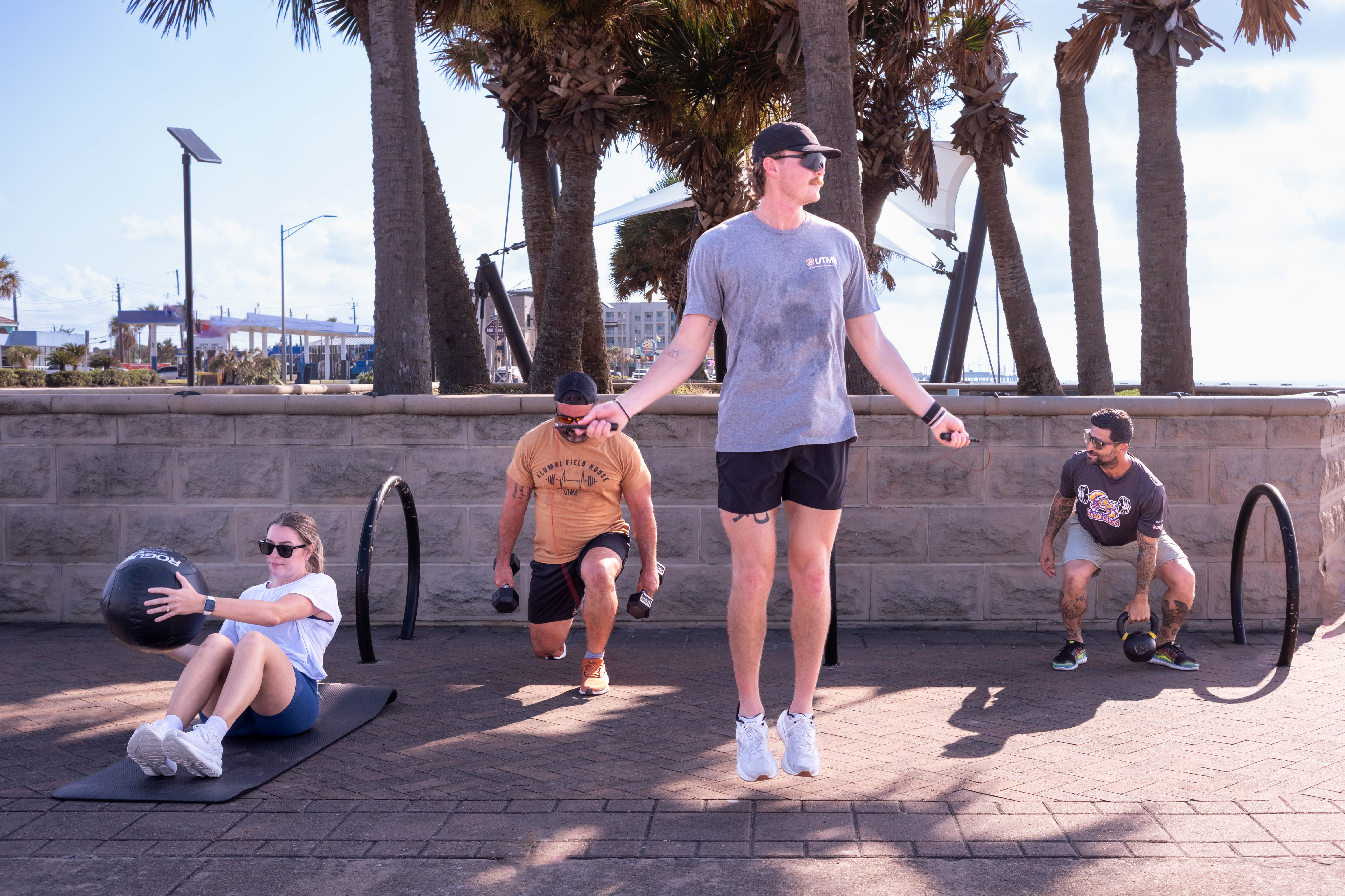 UTMB offers a Wellness & Performance service through the Department of Rehabilitation Services at the UTMB Alumni Field House, led by Tyler Morrison, Cade Sattelmaier, and David Salles (pictured above). Four people exercise outdoors on a paved area near palm trees, using a jump rope, medicine ball, dumbbells, and kettlebell. One person wears a UTMB T-shirt while participating in the workout.