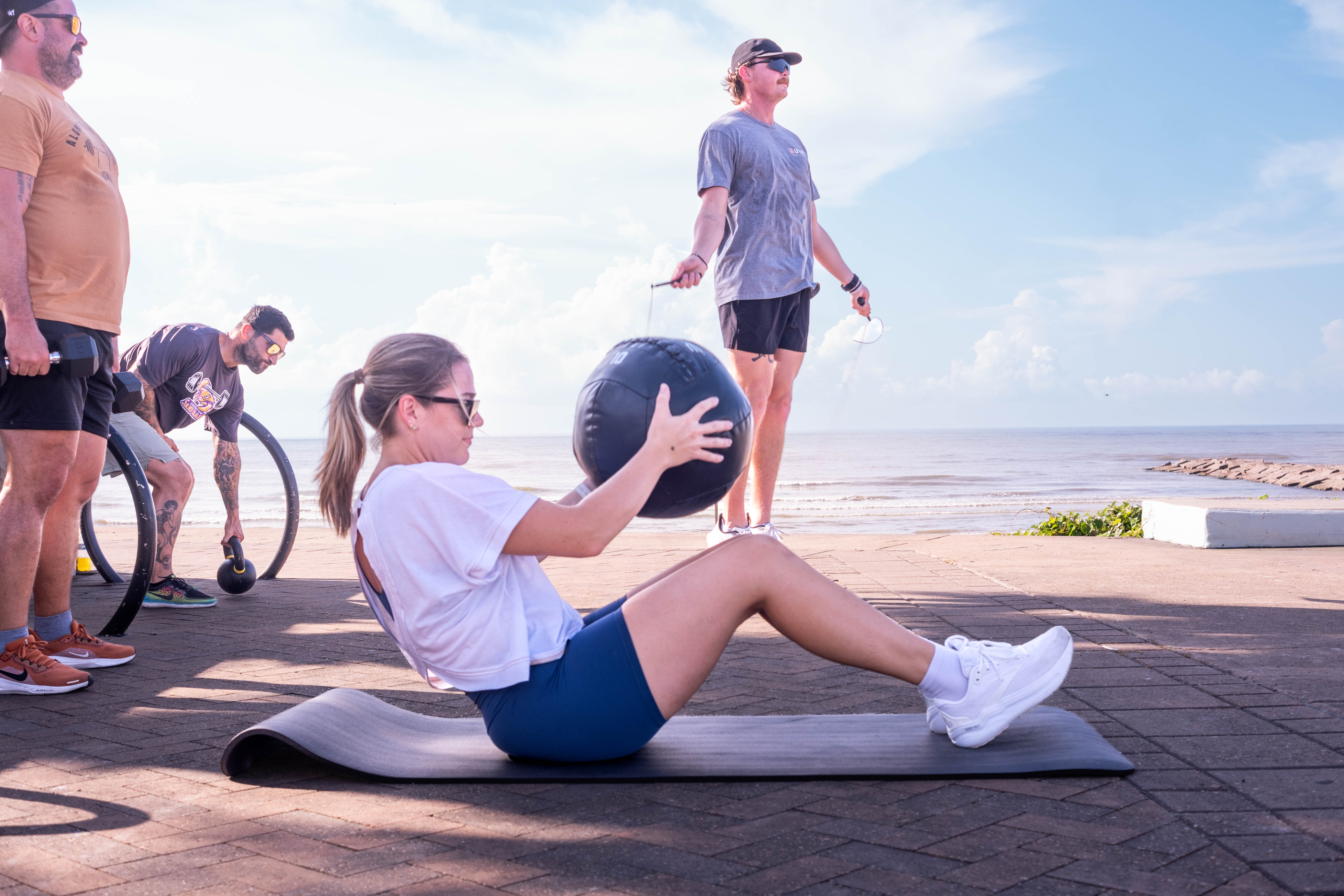 Staying active through regular exercise improves brain, cardiovascular, and metabolic health, all of which contribute to our long-term health. A group exercises outdoors near the shoreline, with one person performing a sit-up while holding a medicine ball and others using a jump rope, dumbbells, and a kettlebell. The ocean and a rock jetty are visible in the background.