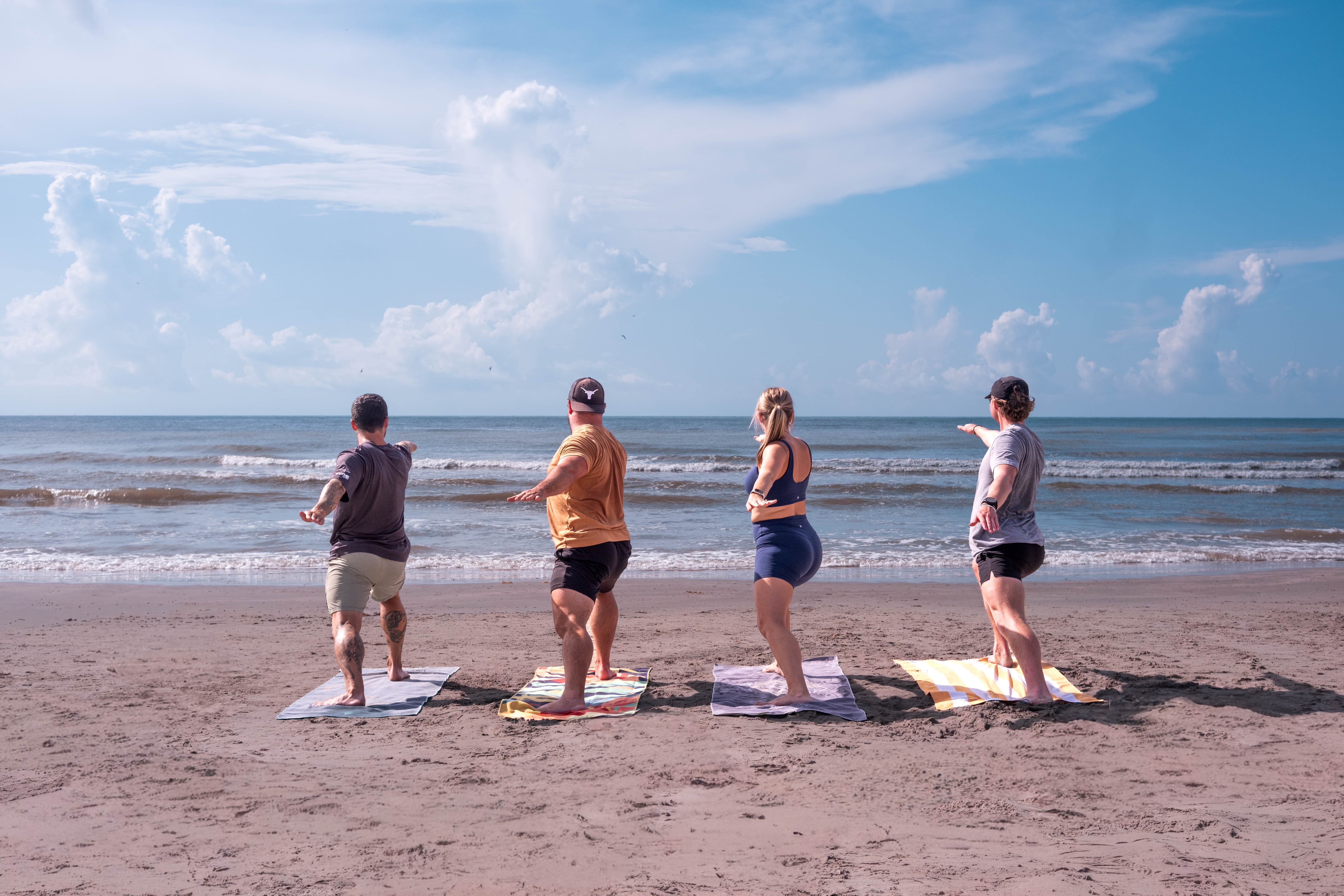 In addition to offering ample opportunity for physical activity, Galveston also offers a beautiful setting for said activity—where better to work out than on the beach? Four people stand on towels at the beach with arms extended, facing the ocean. Gentle waves, sand, and a cloudy sky are visible in the background.