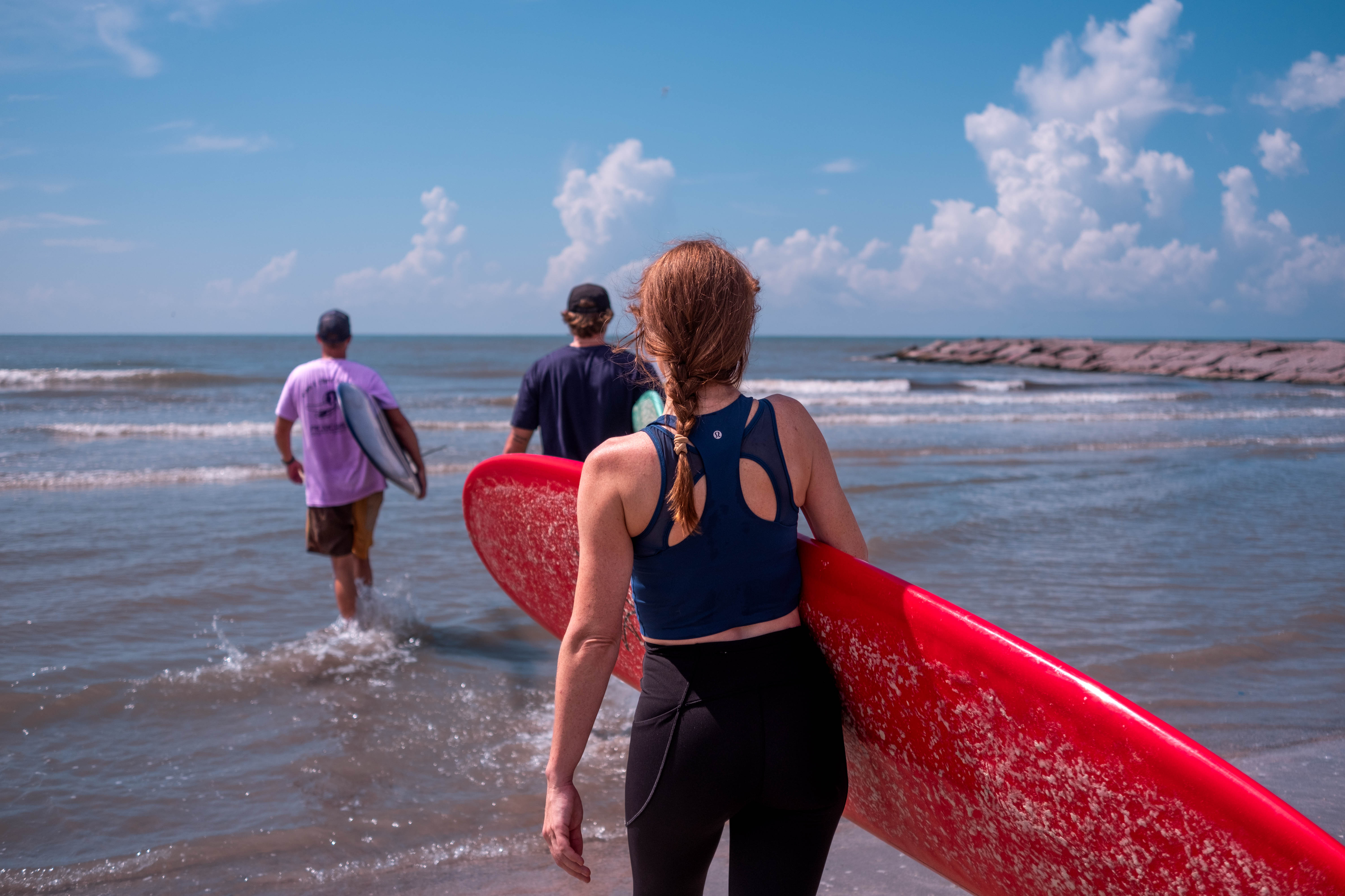 Surfing is known to be beneficial for physical health… Three people carry surfboards into shallow water at the shoreline. Small waves and a rock jetty are visible in the background.