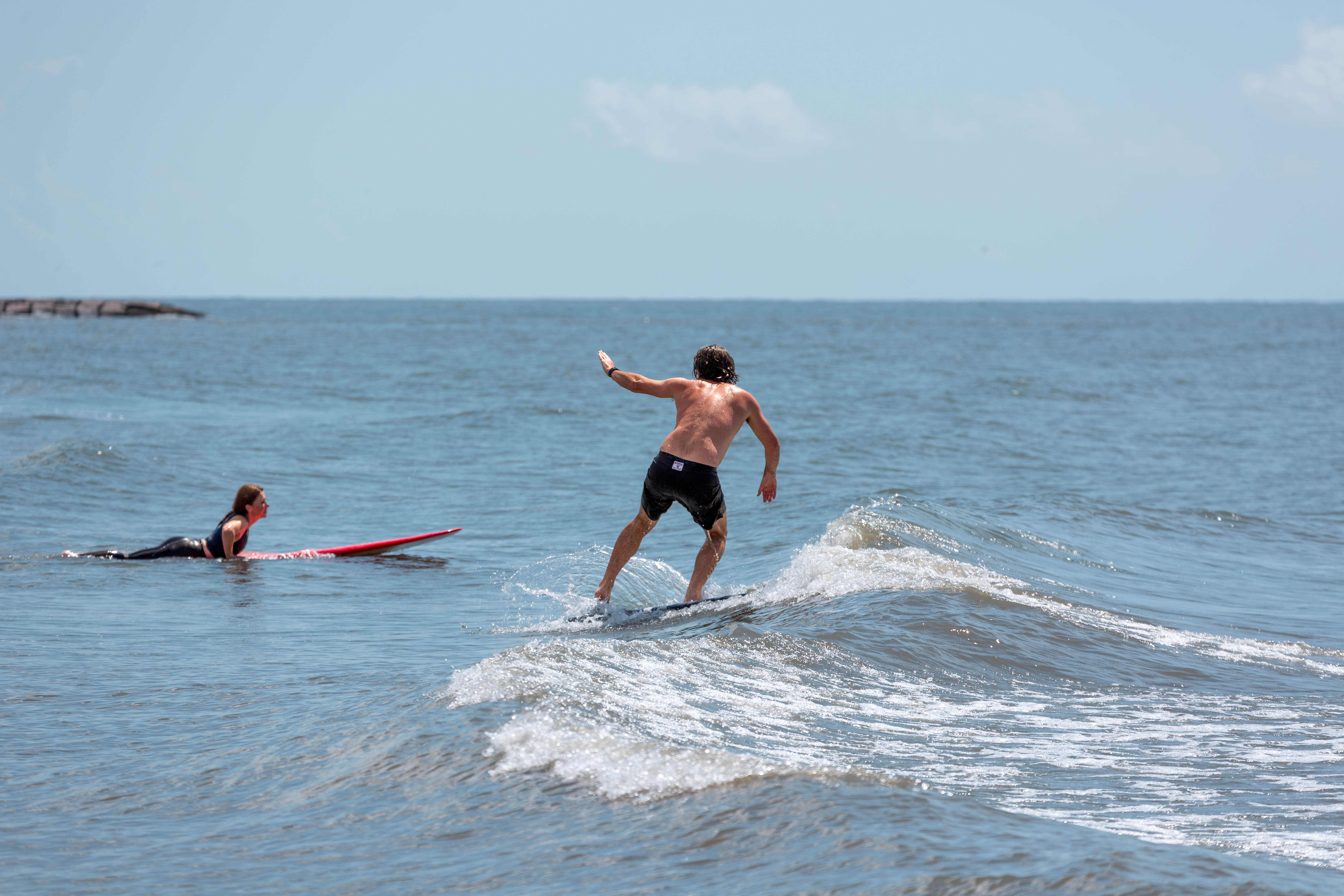 …as well as mental health. One person rides a surfboard on a small wave while another person lies on a red surfboard nearby in the water.