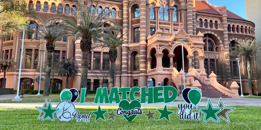 image of "I Matched" Celebratory yard signs in green grass in front of "red" historical building and palm trees on the GAlveston Campus