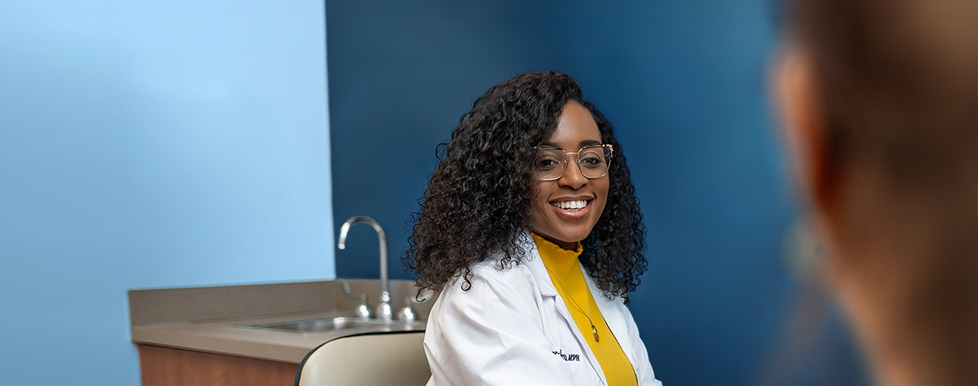 A clinican wearing a white lab coat sits in a clinical exam room with a sink and counter in the background, facing another person whose shoulder is visible in the foreground.