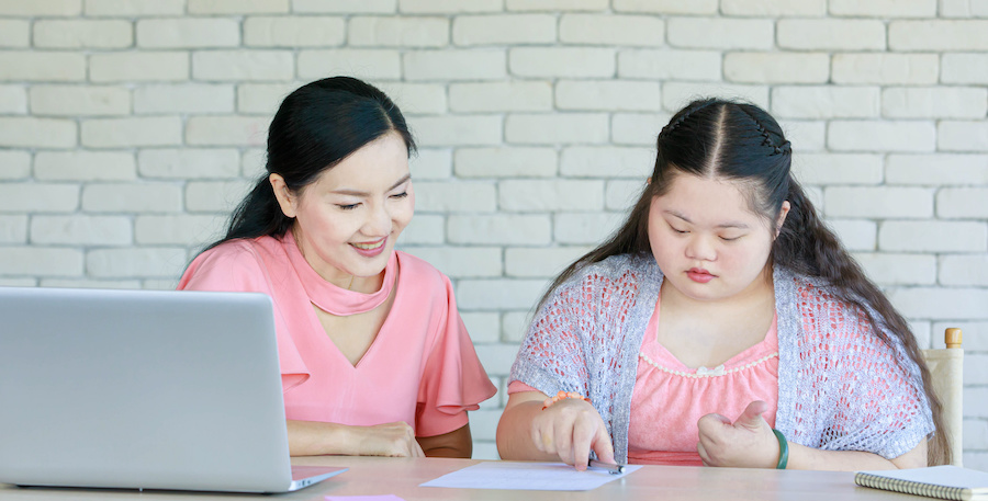 An adult and a child sit at a table with a laptop and papers, reviewing school materials together in a home setting with a brick wall in the background