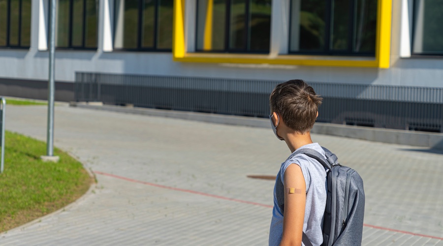 Young boy with gray shirt and gray backpack in front of school with left arm exposed revealing a bandaid on the upper portion of his arm.