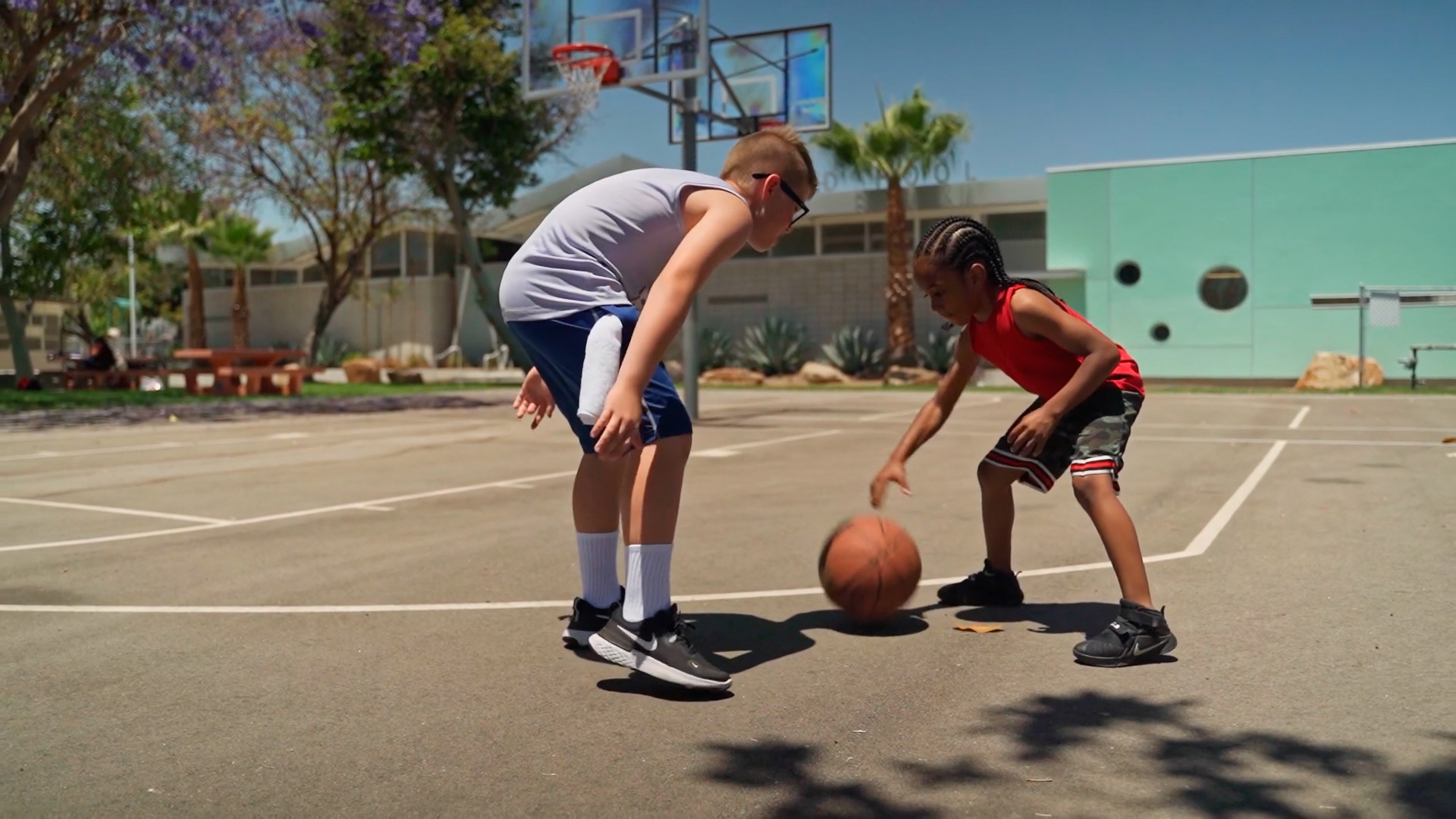 Caucasian boy with glasses playing basketball with younger black boy on an outdoor court in the sun with a palm tree in the background