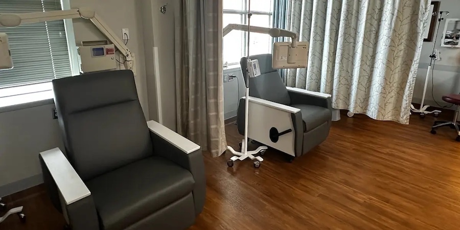 Empty outpatient infusion room with several gray reclining chairs, IV poles, medical equipment and privacy curtains, set on a wood‑tone floor in a clean, clinical environment