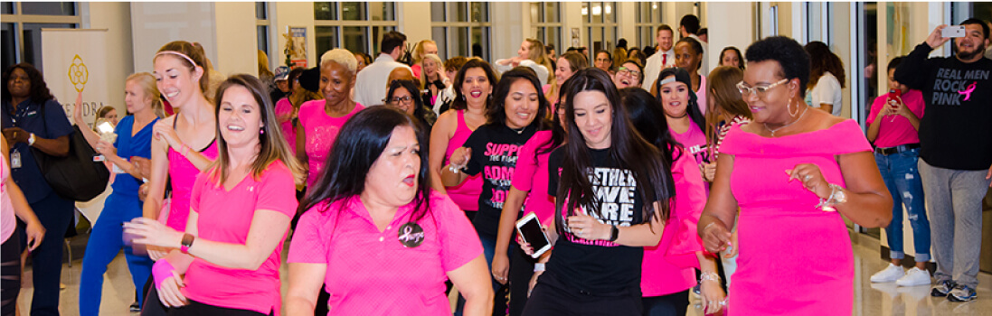 A group of breast cancer survivors and supporters wearing pink gather, smile and dance together at a UTMB Health hospital event celebrating Breast Reconstruction Awareness Day