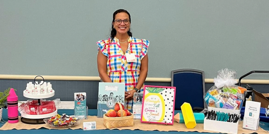A person stands behind a table displaying planners, small gifts, brochures, and baskets at an indoor event. The table is arranged with colorful materials and supplies against a plain wall and chairs in the background.
