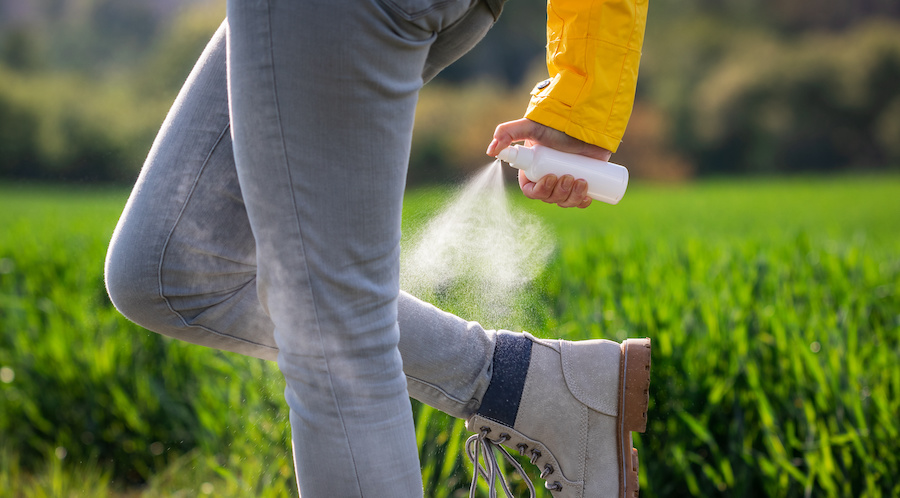 image of someone walking spraying bug spray on their ankles. they have gray denim pants on with ankle high hiking boots.