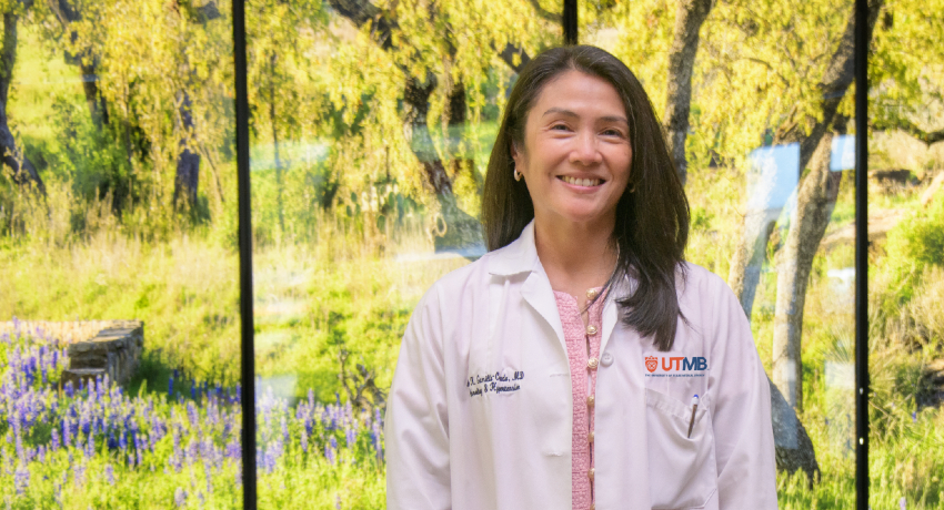 A woman wearing a white UTMB lab coat stands indoors in front of large bright mural featuring trees and purple wildflowers.