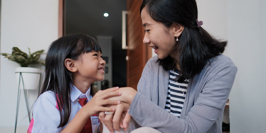 Child wearing a school uniform and backpack sitting to the left of her mother. She is holding her mother's hand and they are conversing and smiling looking at each other