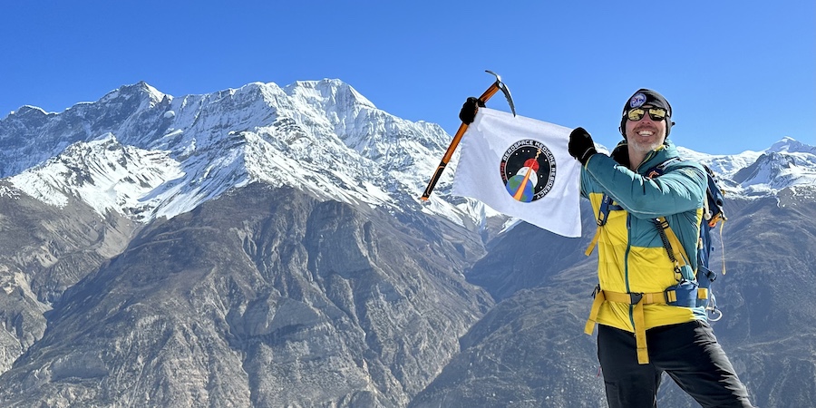 A person in climbing gear stands on a rocky ridge holding a flag with a circular emblem, with snow-covered mountains and a clear blue sky in the background.