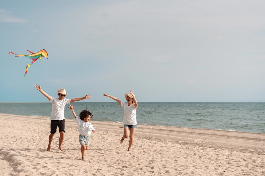 Three people run along a sandy beach flying a colorful kite, with the ocean and calm sky in the background on a sunny day