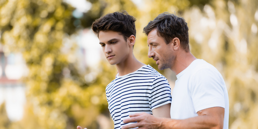 adolescent boy walking with father in front of trees. the boy has on a white shirt with horizontal navy stripes and the dad is wearing a solid white tee. They have serious, concerned expressions and both are brunettes.