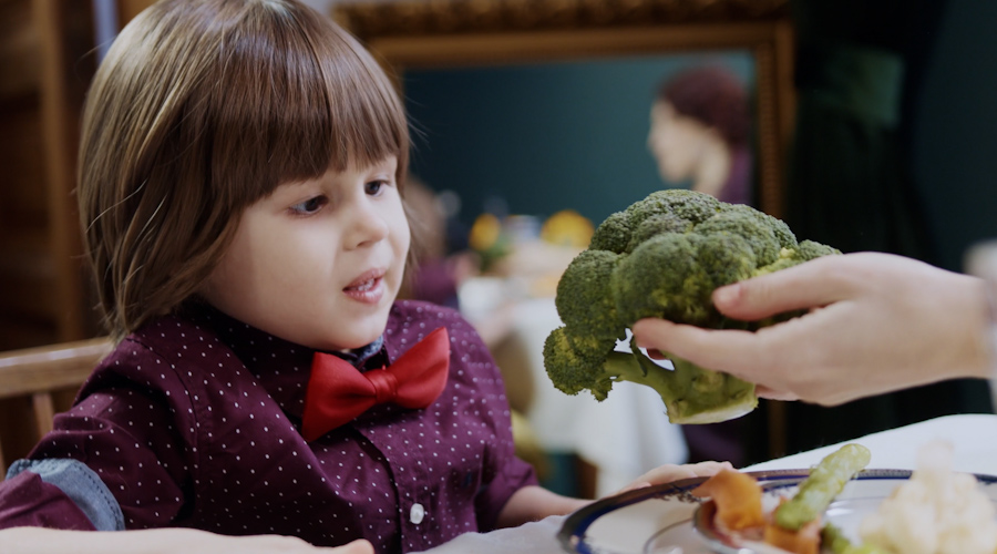 Young child seated at a table looks at a piece of broccoli being offered by an adult hand, with a plate of food in front of the child in an indoor dining setting