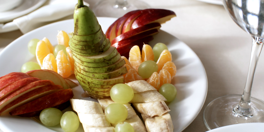 Plate of fresh fruit including sliced apples, pears, bananas, grapes, and orange segments arranged neatly on a white plate beside a glass of water