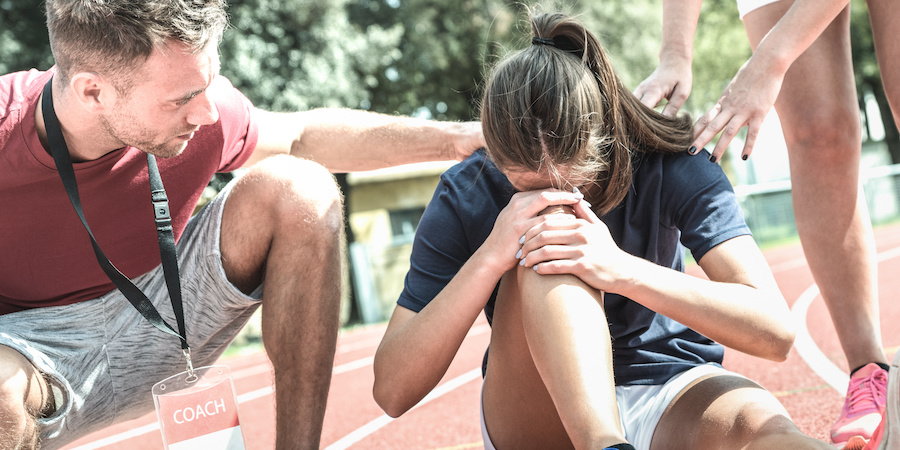 male coach in red shirt and gray shorts consoling injured female athlete sitting on track gripping knee