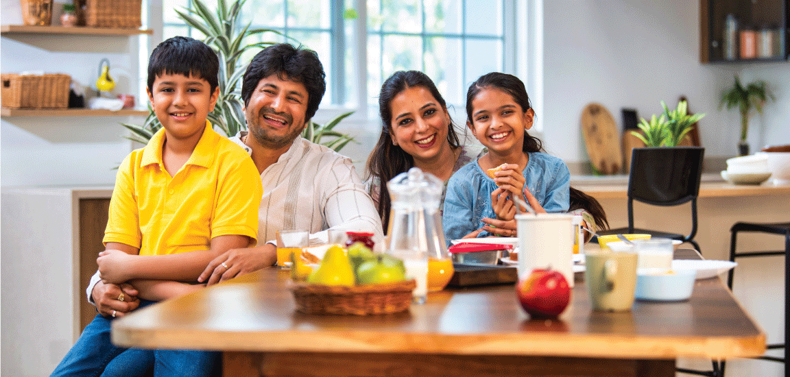 Family seated together at a dining table in a bright kitchen, with adults and children smiling toward the camera and bowls of fruit, drinks, and breakfast items arranged on the table, illustrating healthy eating and family mealtime