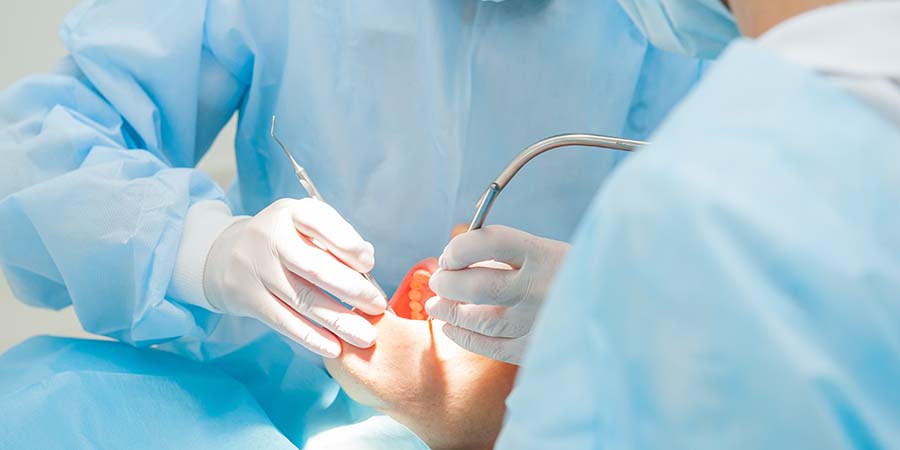 Close-up view of an oral surgery procedure in progress, with gloved hands using surgical instruments inside a patient’s mouth while wearing protective blue surgical gowns in a clinical operating setting
