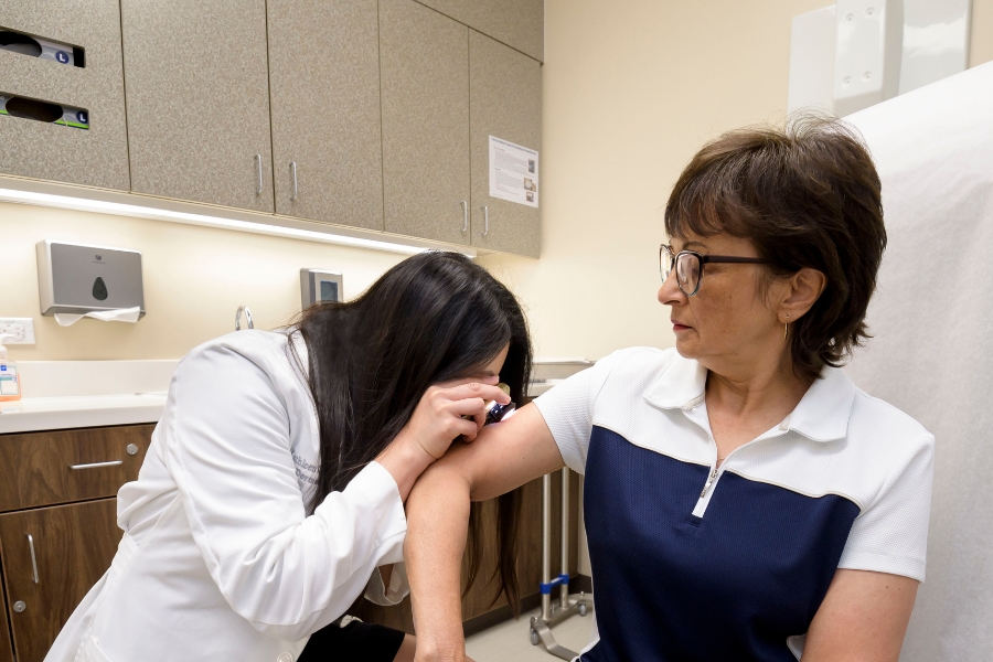 A physician wearing a white coat examines a patient’s upper arm with a handheld magnifying tool in a medical exam room, while the patient sits on an exam table with cabinets and medical equipment in the background