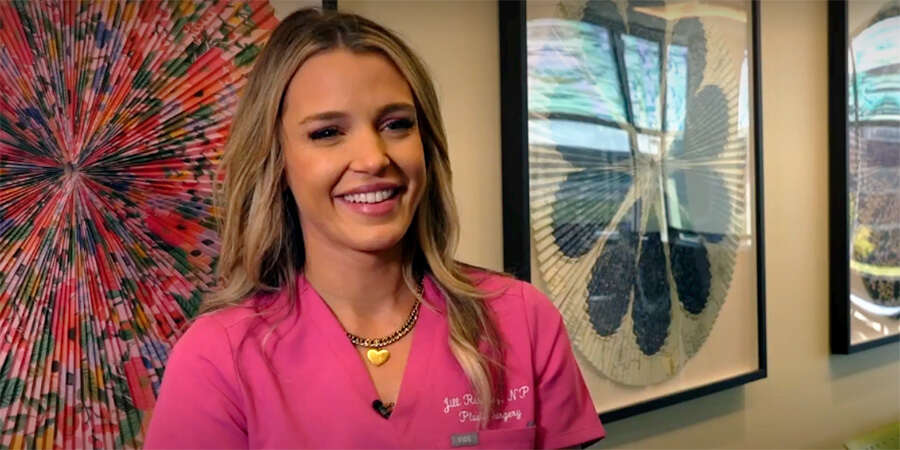 A health care professional wearing a pink scrub top stands indoors in front of framed artwork hanging on a wall