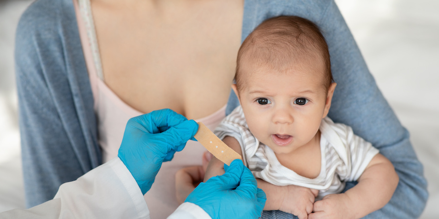A baby sits on an adult’s lap while gloved hands nearby prepare a small adhesive bandage, representing infant immunization and pediatric preventive care