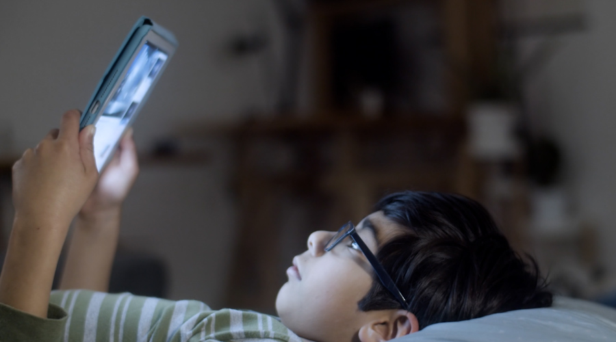 boy with glasses lying in bed holding a tablet in front of his face