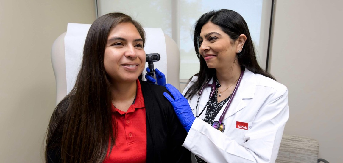 Health care provider uses an otoscope to examine a seated patient’s ear in a medical exam room, with an exam chair, clinical lighting and neutral-colored walls visible in the background