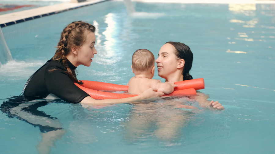 Two adults assist an infant in a swimming pool, using orange flotation noodles to support the child during a supervised water activity