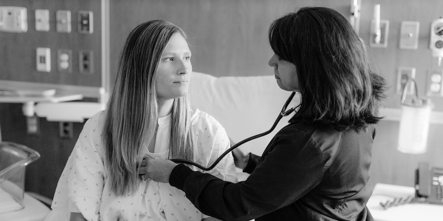 patient wearing gown seated while a physician uses a stethoscope during a check up. The image is in black and white