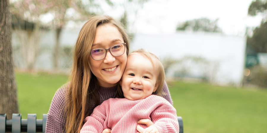 Image of UTMB Health women's health, orthopedics & genetics patient Audrey Solomon, sitting on a bench, wearing glasses & a mauve-colored knit sweater, holding 18-month-old daughter Maisie Solomon. She has on a pink-colored knit sweater both are smiling