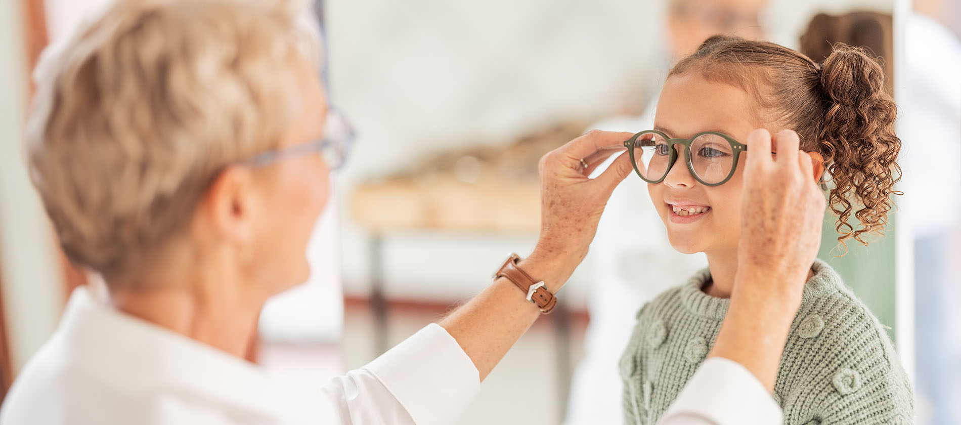 A smiling young girl tries on glasses with the help of an eye care professional during a pediatric vision exam.