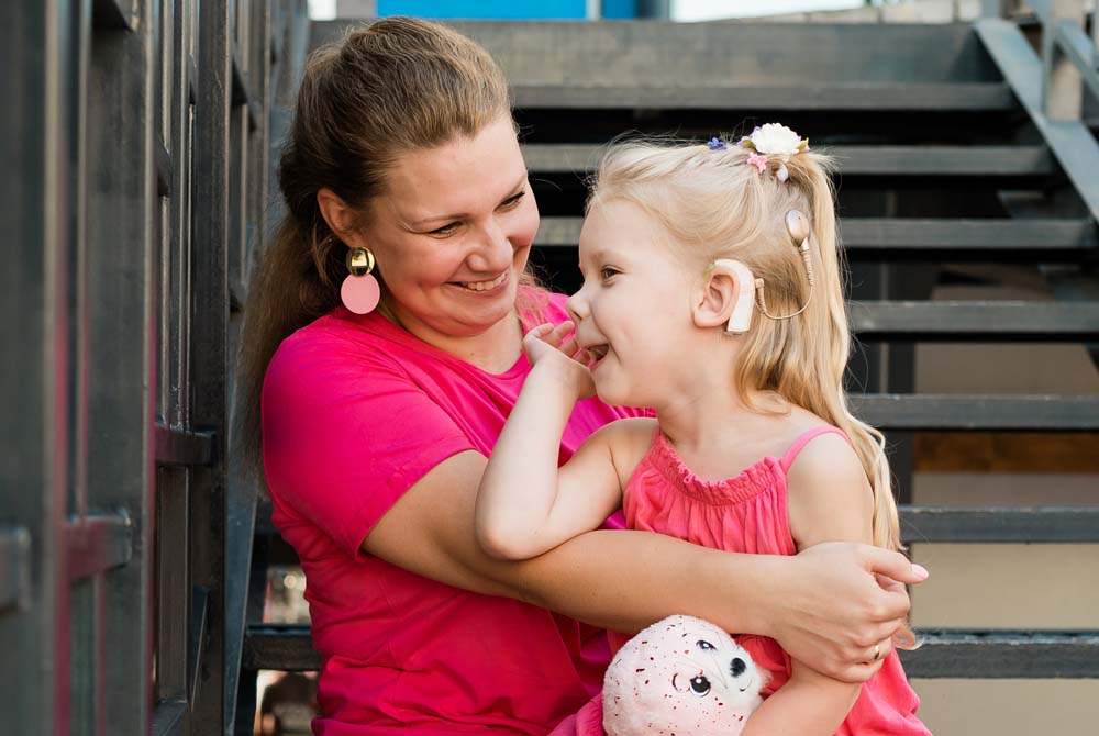 A smiling young girl with a cochlear implant sits in her mother’s lap while they play together outdoors.
