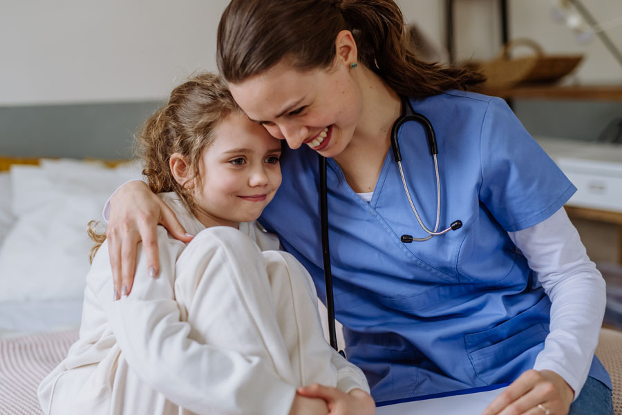 A female nurse wearing scrubs hugs a young child, both smiling while sitting on a hospital bed.