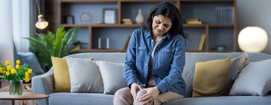 A young woman sits on the couch at home and holds her leg, feeling severe pain in her knee and bones.
