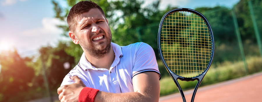 Man grimacing in pain as he touches his injured shoulder while playing tennis outdoors, holding racket in other hand